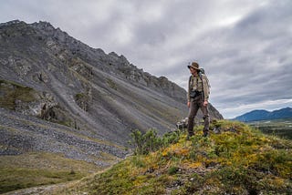 A uniformed biologist stands on a mossy hill with mountains in the background.
