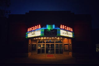 A movie theater after dark, after closing. The neon sign advertising the feature films.