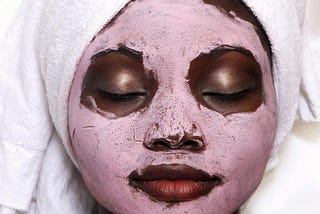 A photo of a beautiful black woman with a pink cosmetic mask on her face.
