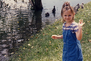 A young girl waves alongside the edge of a lake, with ducks in the distance.