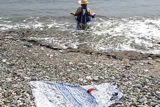 A woman sits in a chair, in surf, facing the ocean. Her blue patterned shift is spread on the pebbled beach in the foreground.