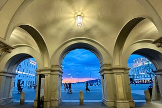 Photo of Trieste’s Piazza dell’Unita d’Italia from behind a tri-arched dome, at sunset with a pink & blue sky in the background.