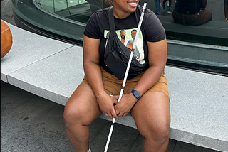 Jasmine Simmons, a Black woman, sitting on a bench, looking to the side and holding a white cane. Courtesy of Jasmine Simmons.