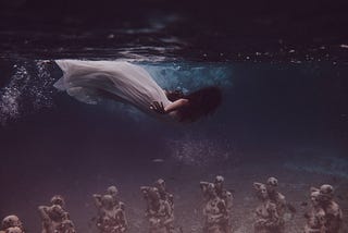 A woman in a white dress, diving close under the water surface, looking down, dark long hair is covering her face. On the ground are mannequins arranged as if they were a couple — but many of them.