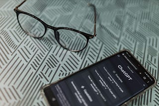 Phone with ChatGPT app on the screen next to a pair of glasses on a patterned table