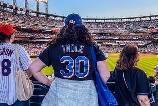 A woman (me) with medium-length brown curly hair, wearing a blue baseball cap and a black t-shirt with “Thole 30” stands facing the baseball diamond at Citi Field