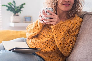 Smiling woman in a yellow sweater holding a coffee mug and reading a book