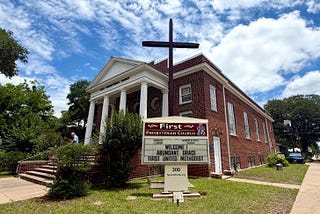 Historic red-brick First Presbyterian Church in Smithville, Texas, with a large cross standing in front beneath a bright blue sky filled with scattered clouds.