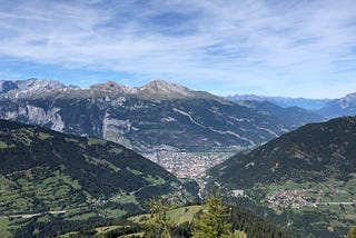 View from the top of a mountain across a valley. In the bottom of the valley there is a very built up town. The lower hills have lots of vegetation and grass, and the tops of the mountains are much more barren and rocky.