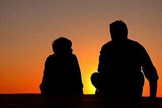 The silhouettes of an adult and a child sitting next to each other against a sunset