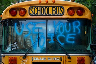 Close up of the front windshield of a big, yellow school bus. On the window, in big, bright, blue spray paint, the words “Use Your Voice” and smaller, in white marker, the words, stacked: “I Love You” “No Justice” “No Peace.” Below the windshield, the bus logo reads, “Blue Bird” in metal letters,  and above the windshield are the words, “School Bus” in black.