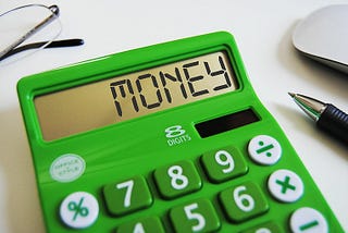 Green calculator on a white background with glasses, an ink pen, and a computer mouse. The calculator screen says “money.”