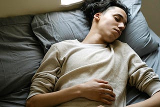 A young person lies on a gray bed with one hand resting on their stomach and eyes closed. They appear calm but slightly tense, as if monitoring their breath or recovering from exhaustion.