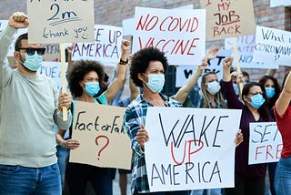 Outdoor protest with a diverse crowd wearing face masks. People hold handmade signs: “WAKE UP AMERICA,” “NO COVID VACCINE,” “I WANT MY JOB BACK,” “2m thank you,” “Fact or Fake?,” and more. Central person with short curly hair raises a white sign. Others lift fists or placards. Brick wall background; atmosphere tense and energetic, with tightly packed participants and varied expressions of concern and determination.