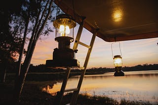 2 lanterns hang by a ladder, in front of a lakeside outdoors while sun is setting.