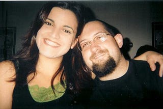 Sherene and Jimbo smiling at one of his parties in 2005. Sherene has long dark hair and is wearing a black top with a green lace neckline. Jimbo has a beard, glasses, and is leaning in with his arm around her. Both look happy and carefree.