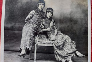 Black and white old photo of two young Chinese women lounge posing in long, floral gowns, 1910s/1920s style bob haircuts.