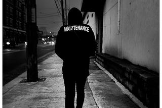 Man walking down the street, shoulders hunched, on his way to work. His jacket says “Maintenance” across the back.