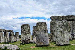Ancient stone pillars and lintels at Stonehenge under a cloudy sky. The weathered stones stand on a grassy plain, with a narrow opening of blue sky visible between layers of grey cloud.