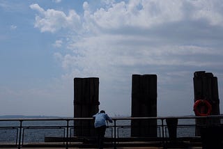 Battery Park, New York City sky and harbor view with man standing alone looking at water.