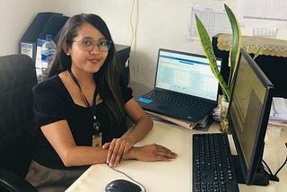 A woman sits at an office desk in front of two computer screens. Her hands rest on the desktop and she is looking straight at the camera.