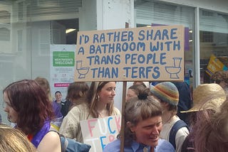 Placard held up above a crowd, saying “I’D RATHER SHARE A BATHROOM WITH TRANS PEOPLE THAN TERFS”.
