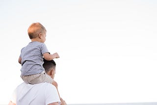Man in white shirt carrying young boy on his shoulders, facing away from the camera, looking into the distance.
