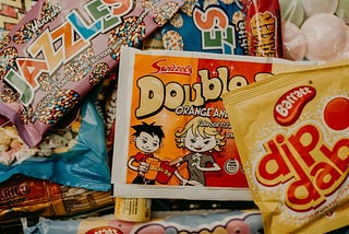 A selection of vintage sweets, including Jazzles, a DibDab, and a sherbert Double Dip. The packaging of the Double Dip depicts a young man offering his candy stick to a young woman, who is reaching for it with a sly grin.