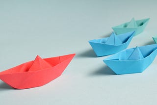 Five origami boats placed on a surface as if racing. The red boat is in the lead, ahead of two blue boats and two green ones.