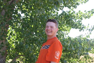 A smiling young man with short hair in an orange baseball shirt standing in front of a tree.