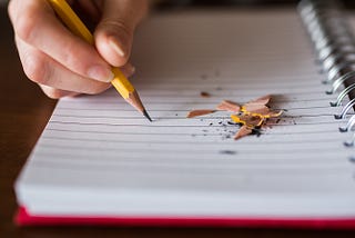 A person is writing in a notebook with a pencil. There are pencil shavings lying on the blank notebook page.