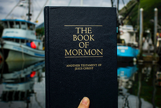 A photo of a copy of The Book of Mormon being held up by a hand against a blurry background on fishing boats in a dock with an overcast blue sky and green hills. The book and hand are in sharp focus and in a different light to the boats, suggesting that the book may be held up to a different photo, or cropped into this one.