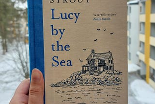 Fariza’s hand holding the copy of Lucy by the Sea with apartment building of grey and yellow paint to the right and snow on the ground and few trees on the left