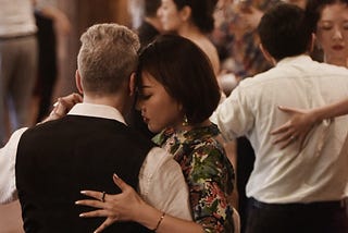 A group of people dance in a room. They are dancing in couples, each couple in a close embrace.