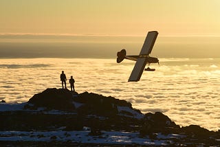 Two people stand on a mountaintop as a single-engine prop plane banks above a sea of clouds…Broken Rainbows by Jim Latham | Photo by Paxson Woelber on Unsplash