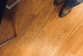 Image of a wood floor with the author’s brown cowboy boots and jeans in the upper right corner, and a part of a chair in the upper left corner.