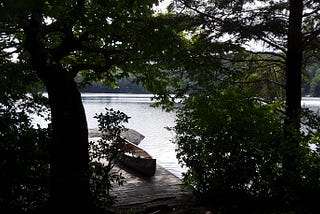 Looking out through trees to a wooden dock with a canoe and a still lake.