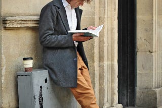 A man standing against a sone wall holding a book in both hands with a coffee at his side.