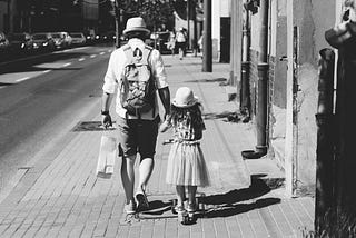 Adult walking on street, holding hand of child who is on scooter.