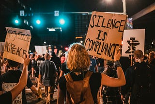 A photo of a group of people protesting in the street at night. A placard that can be seen reads Silence is Compliance