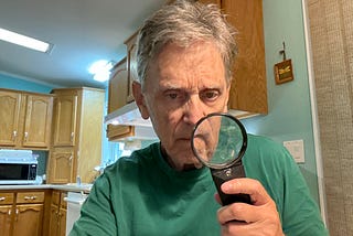 An older man with gray hair and a green shirt sits in a kitchen holding a magnifying glass up to his face, peering through it with a serious expression. Wooden cabinets and a microwave are visible in the background.