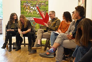 A group of six men and women sit in a semi-circle talking — behind them is a green painting with animal figures and red arrow-shaped sculpture.