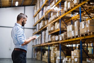 A man wearing a collared shirt and jeans looks at a document with shelves of boxes in the background.