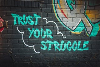 ‘Trust Your Struggle’ graffitied in bright green on to a painted black brick wall