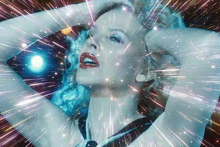 A screen cap from Kylie’s Minogue’s video for “Say Something” showing her with her hands in her hair, looking upward as she appears to be floating through a worm hole in space.