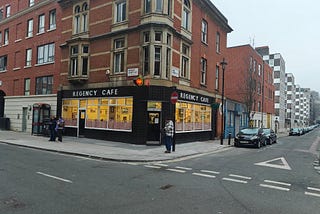 The Regency Cafe is on the corner of a brick building with tall windows. The Cafe has black tiles on the outside with Regency cafe in white capital letters on both sides of the corner building. Inside the cafe the lighting makes it look yellow.