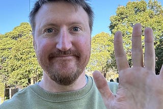 A 40 year old male outside smiling and waving to the camera.