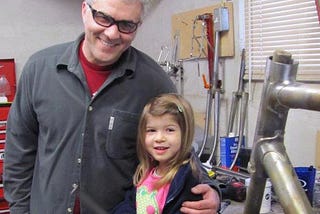 A grinning man with white hair and glasses stands on the left, with his left arm around the shoulder of a smiling young girl. To the right is part of a bike frame.