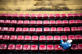 Photo by Bertrand Gabioud on Unsplash // A man sits alone in a stadium of empty red seats.