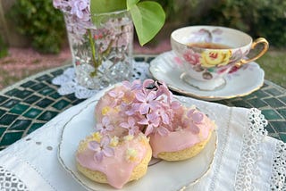 A small garden table with a floral teacup, and three lemon lilac mini donuts on a plate.